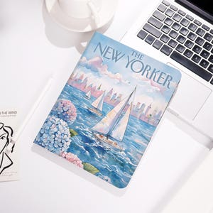 May include: A notebook with a watercolor illustration of sailboats on the water, with the text "The New Yorker" on the cover. The image features blue and pink hydrangeas and a city skyline in the background.