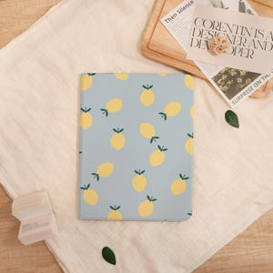 May include: A light blue notebook with a pattern of yellow lemons and green leaves. The notebook is lying on a white cloth. The text "CORENTIN IS A DESIGNER AND DEVELOPER" is visible in the background.