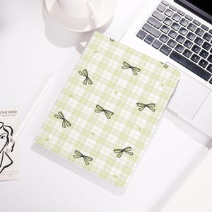 May include: A notebook cover featuring a light green and white plaid pattern, adorned with black bow and heart motifs. The notebook is open on a white surface, beside a laptop and a coffee cup.