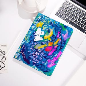 May include: A blue and green watercolor painting of fish, bubbles, and two white rabbits. The fish are yellow, pink, and purple. The background is a blue and green watercolor wash.
