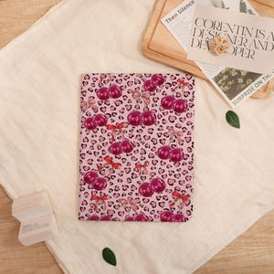 May include: A pink tablet case with a repeating pattern of cherries and leopard print. The cherries are a deep pink with red bows. The background is a light pink with black leopard spots.