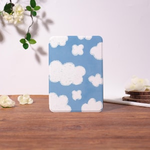 May include: A rectangular, light blue case with a white cloud pattern. The case is standing upright on a wooden surface. White flowers and greenery are in the background.