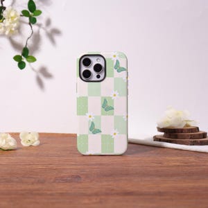 May include: A phone case with a green and white checkered pattern, adorned with illustrations of butterflies and daisies. The case has a smooth finish and a protective design, suitable for a smartphone.