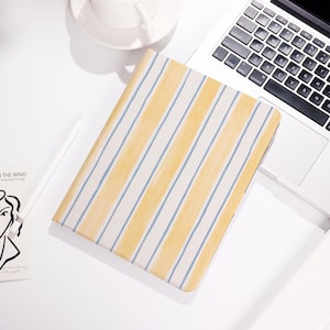May include: A rectangular notebook with a yellow and blue striped pattern. The notebook is lying on a white surface next to a laptop and a white coffee cup. A white stylus and a small art print are also visible.
