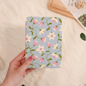 May include: A floral-patterned notebook with a light blue background. The design features pink and cream-colored flowers with green stems and leaves. The notebook is held by a person.