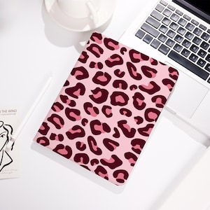 Könnte beinhalten: Rosa Tablet-Hülle mit Leopardenmuster in Rosa- und Burgundertönen. Die rechteckige Hülle liegt auf einer weißen Oberfläche neben einem Laptop und einer Tasse.