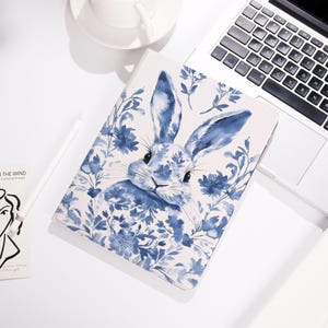 May include: A white notebook with a blue watercolor illustration of a rabbit surrounded by blue floral elements. The notebook is open on a white surface next to a laptop and a coffee cup.
