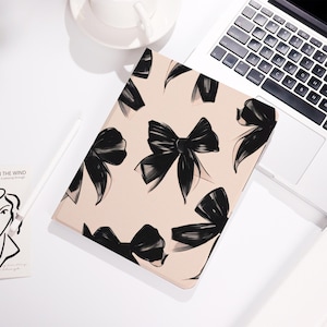 Peut inclure: Une housse de tablette avec un fond rose clair et un motif de nœuds noirs. L'étui rectangulaire est posé sur une surface blanche, à côté d'un ordinateur portable et d'une tasse.