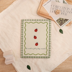 May include: A notebook with a cream-colored background featuring three red ladybugs with black spots. The notebook has a green gingham border and a wavy green inner border. The text "the world is full of tiny wonders" is at the bottom.