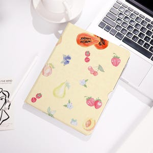 May include: A yellow notebook with a watercolor fruit pattern. The design includes papaya, strawberries, cherries, blueberries, pears, and peaches. The notebook is open, revealing blank pages.
