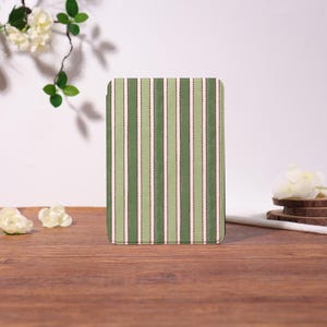 May include: A rectangular case featuring a vertical striped design in varying shades of green, white, and dotted brown. The case is placed on a wooden surface, with white flowers and greenery in the background.