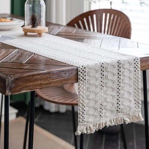 Könnte beinhalten: Ein weißer gehäkelter Tischläufer mit Fransen an den Rändern auf einem Holztisch. Der Läufer hat ein geometrisches Muster und ist perfekt, um jedem Esstisch einen Hauch von rustikalem Charme zu verleihen.