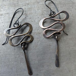 May include: A pair of handcrafted, abstract earrings. Each earring features a looping, wavy design in a bronze-toned metal, with a slender, elongated pendant hanging below. The ear wires are a darker, contrasting colour.