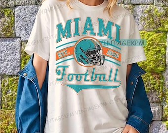 Könnte beinhalten: Weißes T-Shirt mit einem teal- und orangefarbenen Grafikdesign. Das Design enthält das Wort "MIAMI" über einem teal-farbenen Football-Helm, den Text "EST. 1966" und das Wort "Football". Das Shirt wird von einer Person getragen.
