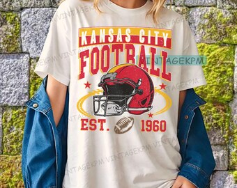 Op de afbeelding: Wit T-shirt met een rode en gele afbeelding met de tekst "Kansas City Football" boven een footballhelm en -bal. Het shirt bevat ook de tekst "EST. 1960".