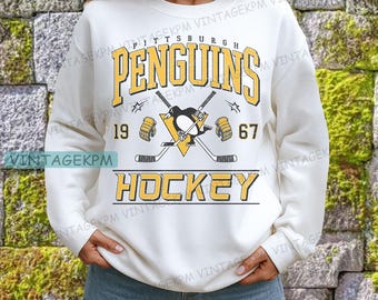 May include: Cream-colored sweatshirt featuring the text "Pittsburgh Penguins Hockey" in yellow and black. The design includes crossed hockey sticks, a penguin logo, and the numbers "19" and "67".