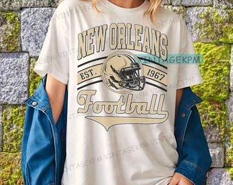 May include: Cream-colored t-shirt with a football-themed graphic. The design features the words "New Orleans Football" in a vintage style, along with a football helmet and the year "1967". The shirt is worn with a blue denim jacket.