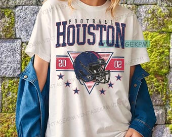 Op de afbeelding: Wit T-shirt met een voetbalthema. Het ontwerp bevat de woorden "FOOTBALL HOUSTON" in blauwe en rode letters, een blauwe voetbalhelm, de cijfers "20" en "02" en blauwe sterren. Het shirt wordt door een persoon gedragen.
