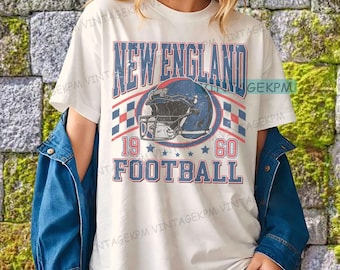 以下が含まれることがあります： クリーム色のTシャツ。青と赤で「NEW ENGLAND FOOTBALL 1960」の文字とフットボールヘルメット、星が描かれたヴィンテージ風のデザイン。レトロなスポーツテーマ。
