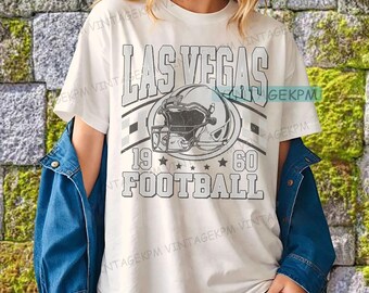 以下が含まれることがあります： 「LAS VEGAS FOOTBALL」と書かれたヴィンテージスタイルのグラフィックデザインが特徴の白いTシャツ。デザインにはフットボールのヘルメット、数字の「19」と「60」、星が含まれています。Tシャツは人が着ています。