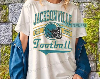 May include: A white t-shirt with the words "Jacksonville Football" in teal and yellow, and a teal football helmet graphic. The shirt also has the text "EST. 1995". The person is wearing a blue denim jacket.