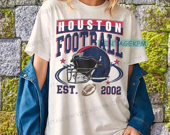Op de afbeelding: Wit T-shirt met een Houston Football-afbeelding. Het ontwerp toont een blauw-rode voetbalhelm, een voetbal en de tekst "HOUSTON FOOTBALL EST. 2002". Het shirt heeft een vintage, distressed look.