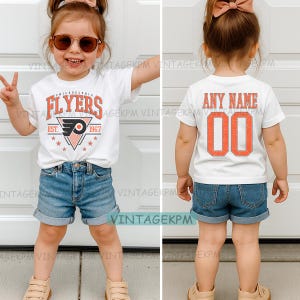 May include: A white t-shirt with "FLYERS" in orange lettering and a team logo. The shirt also has the text "EST. 1967" and stars. The child is wearing denim shorts. The back of the shirt says "ANY NAME" and "00".