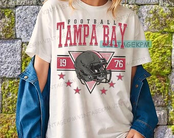Puede incluir: Camiseta blanca con un diseño gráfico rojo y negro. El diseño incluye las palabras "FOOTBALL TAMPA BAY", un casco de fútbol, los números "19" y "76" y estrellas rojas. La camiseta la lleva una persona.