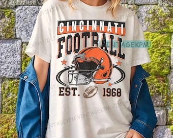 Può includere: T-shirt bianca con un design Cincinnati Football. Il design include un casco da football arancione, il testo "CINCINNATI FOOTBALL" in nero e arancione e l'anno "1968".
