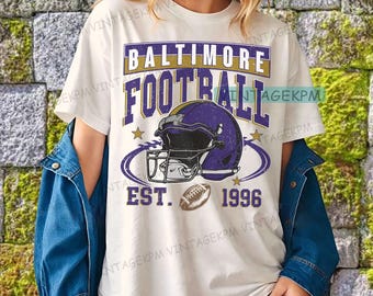 Puede incluir: Camiseta blanca con un gráfico morado y dorado que dice "BALTIMORE FOOTBALL EST. 1996". El gráfico incluye un casco de fútbol americano y un balón. La camiseta la lleva una persona.