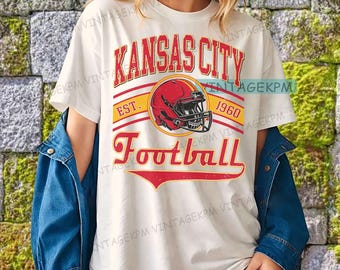 Könnte beinhalten: Cremefarbenes T-Shirt mit dem Text "Kansas City Football" in Rot und Gelb, mit einer Footballhelm-Grafik. Das Shirt enthält den Text "Est. 1960". Das Shirt wird mit einer blauen Jeansjacke getragen.