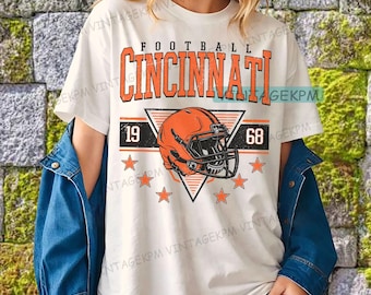 以下が含まれることがあります： ヴィンテージフットボールのデザインが施された白いTシャツ。オレンジ色の「Cincinnati」の文字、フットボールのヘルメット、数字の「19」と「68」、オレンジ色の星がデザインされています。青いデニムジャケットがTシャツの上に着用されています。