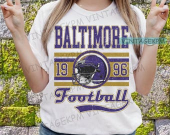 Può includere: Maglietta bianca con un design vintage che presenta il testo "Baltimore Football" con un'immagine di un casco da football viola e oro e i numeri "19" e "96" sui lati.