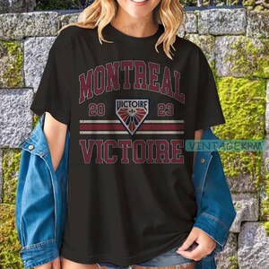 May include: Black t-shirt with the word "MONTREAL" in maroon, above "20 VICTOIRE 23" and "VICTOIRE" in maroon. The shirt has a diamond-shaped emblem with a fleur-de-lis design. The shirt is worn with a denim jacket.