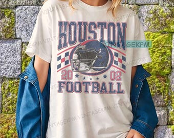 Op de afbeelding: Crèmekleurig T-shirt met een distressed grafisch ontwerp. Het ontwerp toont het woord "HOUSTON" in blauw en rood, een footballhelm, de cijfers "20 02" en het woord "FOOTBALL". Het shirt heeft een klassieke ronde hals en korte mouwen.