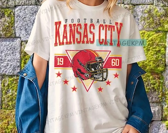 Könnte beinhalten: Cremefarbenes T-Shirt mit einem rot-gelben Grafikdesign. Das Design zeigt die Worte "FOOTBALL KANSAS CITY" in Rot, einen roten Footballhelm, die Zahlen "19" und "60" sowie rote Sterne. Das T-Shirt wird von einer Person getragen.