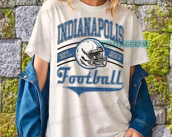 May include: White t-shirt with a blue and black Indianapolis Football graphic. The design includes a football helmet, the text "INDIANAPOLIS", "EST. 1953", and the word "Football". The shirt is worn with a blue denim jacket.