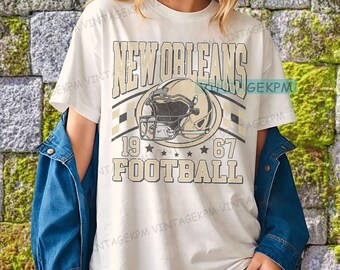 May include: Cream-colored t-shirt featuring the words "NEW ORLEANS" above a football helmet graphic. Below the helmet are the numbers "19" and "67" with the word "FOOTBALL" underneath. The design is in beige and black.