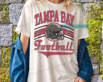 Puede incluir: Camiseta blanca con un gráfico rojo y negro. El gráfico incluye las palabras "TAMPA BAY FOOTBALL" con un casco de fútbol americano y el año "1976". La camiseta tiene un aspecto vintage.