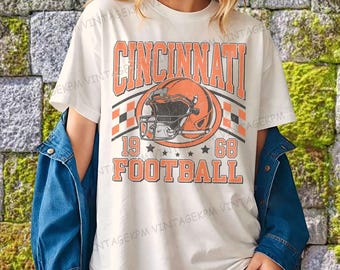 Può includere: T-shirt color crema con la scritta "CINCINNATI" in lettere arancioni a blocchi sopra una grafica di casco da football arancione. La maglietta mostra anche "19 68 FOOTBALL" in arancione. La maglietta è indossata con una giacca di jeans blu.