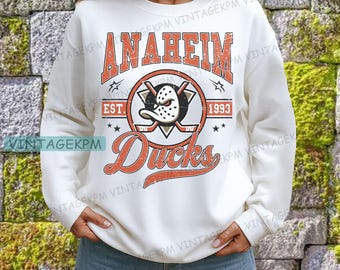 May include: A white sweatshirt featuring the Anaheim Ducks logo in orange and black. The design includes the team name, a hockey mask, crossed hockey sticks, and the text "EST. 1993". The word "Ducks" is written in a cursive font.