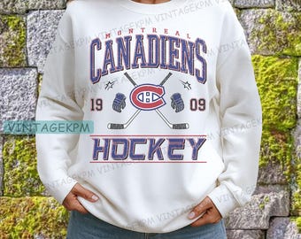 Puede incluir: Sudadera de color crema con el texto "MONTREAL CANADIENS" en azul y rojo, con palos de hockey cruzados y los años "19" y "09". La palabra "HOCKEY" también se muestra en azul y rojo.