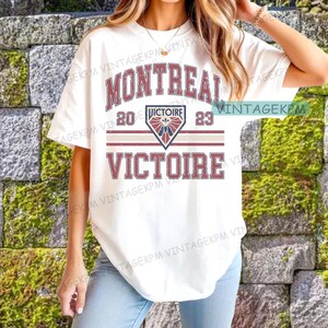May include: White t-shirt featuring the word "MONTREAL" in maroon, above "20 VICTOIRE 23" and "VICTOIRE" in maroon. The design includes a shield graphic with a red, white, and blue design. The shirt has a vintage aesthetic.