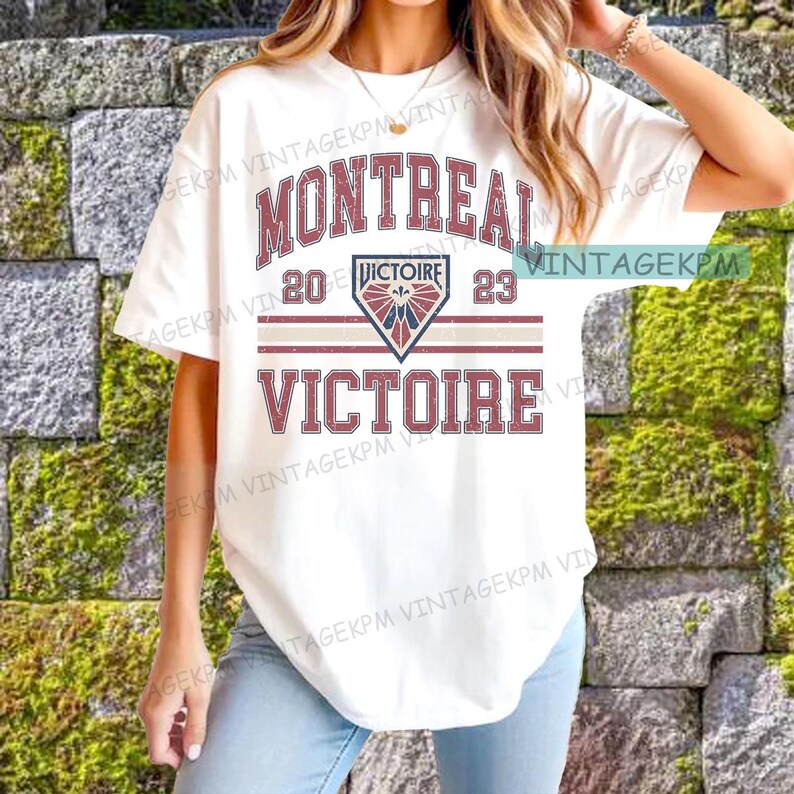 May include: White t-shirt featuring the word "MONTREAL" in maroon, above "20 VICTOIRE 23" and "VICTOIRE" in maroon. The design includes a shield graphic with a red, white, and blue design. The shirt has a vintage aesthetic.