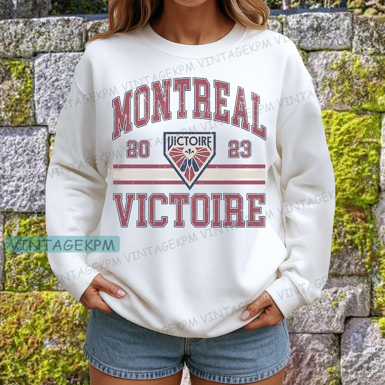 May include: Cream-colored sweatshirt with the word "MONTREAL" in maroon arched lettering. Below, the text "VICTOIRE" is displayed. A crest with the text "VICTOIRE" and the numbers "20" and "23" are also visible. The sweatshirt has a crew neck.