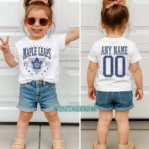 May include: A white t-shirt with a Toronto Maple Leafs logo on the front and the text "ANY NAME" and the number "00" on the back. The child is also wearing denim shorts.