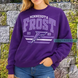 May include: A purple sweatshirt with the text "MINNESOTA FROST" in a light purple, collegiate-style font. Below the text is a stylized "F" logo with the numbers "20" and "23" on either side. The sweatshirt is a solid purple color.