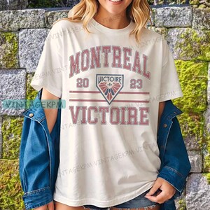 May include: Cream-colored t-shirt with the word "MONTREAL" in red, arched across the top. Below, a graphic with "20" and "23" on either side of a shield. The word "VICTOIRE" is printed below in red.