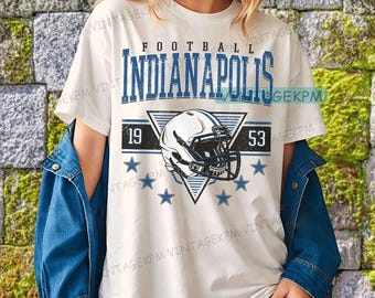 May include: Cream-coloured t-shirt featuring a blue and white graphic. The design displays the word "FOOTBALL" above "INDIANAPOLIS" in blue block letters. A football helmet is in a triangle, with the numbers "19" and "53" and blue stars below.