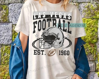 以下が含まれることがあります： 「LAS VEGAS FOOTBALL」の文字とフットボールのヘルメットとフットボールのグラフィックデザインが施された白いTシャツ。「EST. 1960」の文字も含まれています。デザインは黒、グレー、茶色です。